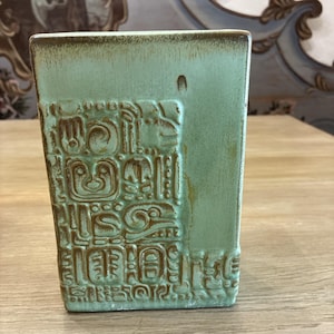 May include: A rectangular ceramic vase with a light green glaze and a brown wash. The front surface features a relief pattern of stylized geometric shapes and symbols, with a smooth, unpatterned vertical section on the right side of the vase.