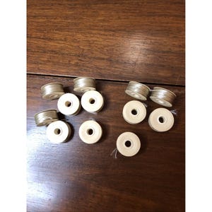 May include: A collection of nine sewing machine bobbins with light beige thread wound around white plastic spools. The bobbins are arranged in two loose groups, showing the circular shape and the central hole of each individual spool component.