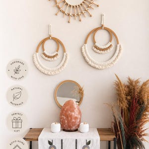 May include: A sunburst mirror, two circular macrame wall hangings with wooden beads, a round mirror, a pink salt lamp, two white textured candle holders, and a white table runner with a floral pattern on a wooden console table with dried grass.