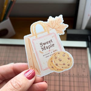 Op de afbeelding: Een gestanste sticker met een beige papieren boodschappentas met de tekst Sweet Maple Bakery & Cafe Made With Love, versierd met een lichtoranje esdoornblad en twee koekjes met zichtbare donkerbruine chocoladestukjes.
