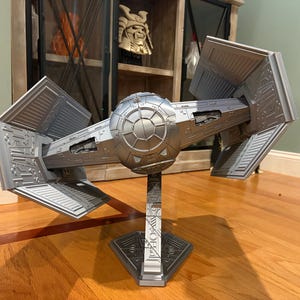 May include: A silver-colored TIE Interceptor model spacecraft featuring a central spherical cockpit and four angled solar array wings. The model is mounted on a vertical display stand with a geometric base, showcasing intricate panel detailing.