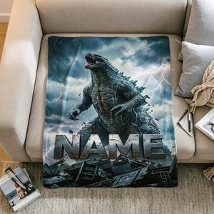 May include: A rectangular throw blanket featuring a large, dark gray, textured monster standing amidst rubble under a stormy sky with lightning bolts. The word NAME is printed in large, metallic, block letters across the lower center section.