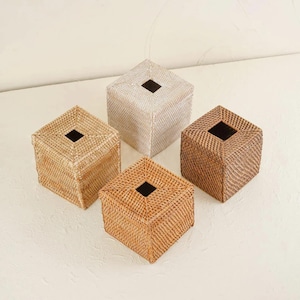 May include: Four square tissue box covers crafted from woven rattan in varying shades of natural brown, tan, and light beige. Each box features a centred square opening at the top, showcasing a detailed, textured weave pattern throughout the exterior.