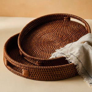 May include: Two round, brown woven rattan trays with integrated handles, stacked one inside the other. A light beige, textured fabric cloth with frayed edges and a thin, dark, wavy stitched pattern is draped over the edge of the trays.