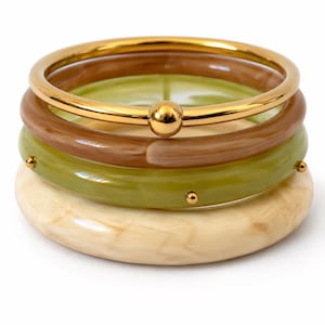 Pode incluir: Uma pilha de quatro pulseiras composta por um anel de metal dourado polido com uma esfera central, um anel de resina castanha translúcida, um anel de resina verde-azeitona com pequenos tachas douradas e um anel de resina creme marmoreado.