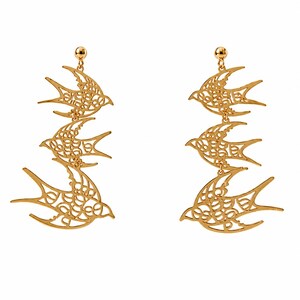Pode incluir: Um par de brincos pendentes em tom dourado com uma disposição vertical de três silhuetas de andorinhas. Cada pássaro apresenta um intrincado padrão de filigrana vazada em todo o corpo e asas, suspenso por um pino simples.