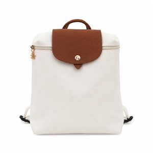 Pode incluir: Uma mochila de lona branca com uma aba de couro castanho texturizado e um fecho de botão de pressão dourado. A mala inclui uma pega superior, um compartimento principal com fecho de correr dourado e alças ajustáveis para os ombros.