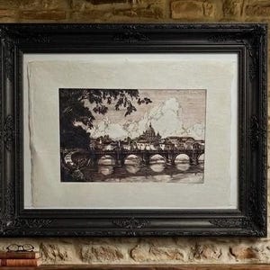 May include: A black ornate frame containing a monochromatic etching of a cityscape featuring a multi-arched bridge over a river, with a large domed building in the background, set against a light-colored textured paper matting.