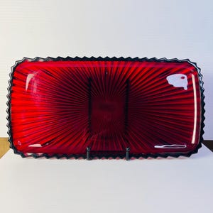 Könnte beinhalten: Ein rechteckiges Tablett aus rotem Glas mit einem strahlenförmigen Muster, das von der Mitte ausgeht. Das Tablett hat einen gezackten Rand und eine glatte, glänzende Oberfläche. Ein kleiner weißer Aufkleber befindet sich in der oberen rechten Ecke.