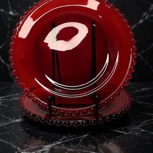 May include: Two round, deep red glass plates featuring a decorative beaded edge. The plates are translucent, allowing light to pass through the glass, and are displayed vertically on a black wire stand to showcase the circular design and texture.
