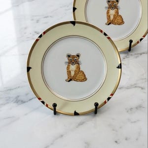 May include: Two round porcelain plates featuring a central illustration of a seated leopard cub. The plates have a cream-colored rim with a gold border and small black and red geometric accents. Each plate is displayed on a black metal stand.
