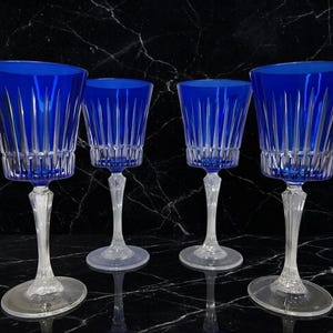 May include: A set of four cobalt blue crystal wine glasses featuring vertical cut patterns on the bowls and clear, faceted stems. Each glass has a wide, circular base and a flared rim, showcasing a deep blue color contrasted with clear glass.
