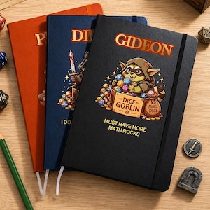 Könnte beinhalten: Drei Notizbücher mit festem Einband in Schwarz, Blau und Orange. Das schwarze Notizbuch zeigt einen Cartoon-Waschbären mit Kapuzenumhang, der bunte Würfel hält, sowie den aufgedruckten Text GIDEON, DICE GOBLIN und MUST HAVE MORE MATH ROCKS.