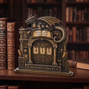 May include: A bronze-toned decorative book tracker featuring a sculpted dragon perched atop an arched library facade. The piece displays the text BOOKS READ THIS YEAR and DRAKOS'S LIBRARY, with three rotating number tiles currently set to 415.