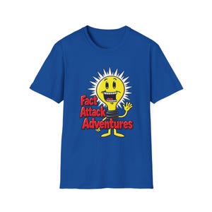 Op de afbeelding: Een koningsblauw t-shirt met korte mouwen en een afbeelding van een geel gloeilampfiguur met een brede glimlach en stekelige stralen. De tekst "Fact Attack Adventures" staat in dikke rode letters links van het figuur.