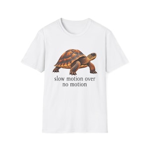 Puede incluir: Una camiseta blanca de manga corta con un gráfico de una tortuga marrón y naranja. Debajo de la tortuga, el texto "slow motion over no motion" aparece en una fuente serif negra. La camiseta tiene un cuello redondo y un diseño estándar.