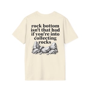 May include: A cream-coloured short-sleeve t-shirt featuring a black graphic print on the back. The text reads, rock bottom isn't that bad if you're into collecting rocks, positioned above a detailed black and white illustration of several rocks.
