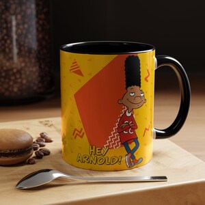 May include: A yellow ceramic mug with a black interior and handle, featuring a graphic of the cartoon character Gerald Johanssen from Hey Arnold! with his tall hair, red shirt, and blue jeans, alongside the text HEY ARNOLD! in yellow letters.