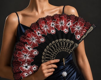 Puede incluir: Un abanico plegable negro con intrincados bordados florales en rojo y blanco a lo largo del borde festoneado. El abanico incluye patrones decorativos dorados cerca de la base, un pequeño dije en forma de flor blanca y una borla negra colgante.