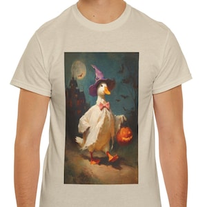 Può includere: Una t-shirt beige con una stampa grafica di un'anatra bianca vestita da fantasma con un cappello da strega viola e un papillon rosa, che trasporta una zucca arancione luminosa in uno scenario notturno scuro e spettrale.