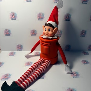 May include: A doll with brown hair, a red and white hat, and red and white striped legs with black shoes, sitting inside a bright orange cylindrical post box. The doll has a white collar and red sleeves with white cuffs, positioned upright.