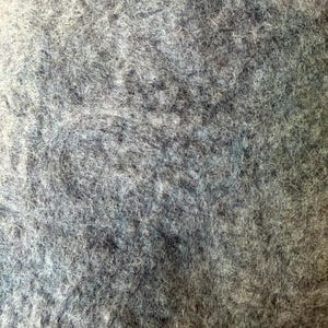 May include: A close-up view of a dense, felted wool textile featuring a mottled blend of charcoal gray, slate blue, and off-white fibers. The surface displays a thick, matted, and fibrous texture with an uneven, organic distribution of colors throughout the material.
