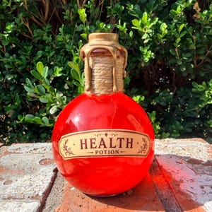 May include: A round glass bottle filled with bright red liquid, featuring a label that reads HEALTH POTION. The neck of the bottle is wrapped in natural twine and topped with a gold-coloured wax seal that drips down the sides of the glass.
