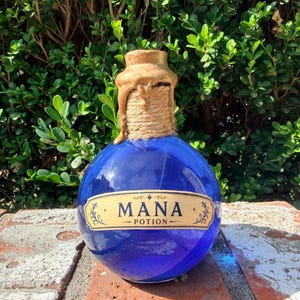 May include: A round, cobalt blue glass bottle featuring a tan label with the text MANA POTION in black lettering. The neck of the bottle is wrapped in natural twine and topped with a metallic gold wax-like seal that drips down the side.