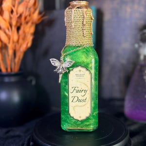 May include: A rectangular glass bottle filled with shimmering green liquid, featuring a gold-colored textured neck with dripping wax detail. A silver-toned fairy charm hangs from a chain, and a label reads Side Quest Potions Fairy Dust.