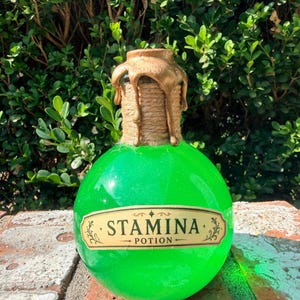 May include: A round glass bottle filled with bright green liquid, featuring a label that reads STAMINA POTION. The neck of the bottle is wrapped in natural twine and topped with metallic gold wax that drips down the sides of the glass container.