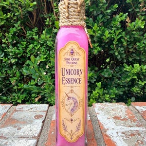 May include: A tall, rectangular glass bottle filled with bright pink liquid. The label reads Side Quest Potions Unicorn Essence with a unicorn illustration. The neck is wrapped in thick twine topped with dripping gold-colored wax and a small charm.