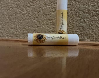 Op de afbeelding: Twee witte cilindrische lippenbalsemhulzen met een geel etiket met een zonnebloemillustratie en de tekst SunnySnoutsStudio. Eén huls staat rechtop en de andere ligt horizontaal ervoor op een reflecterend oppervlak.