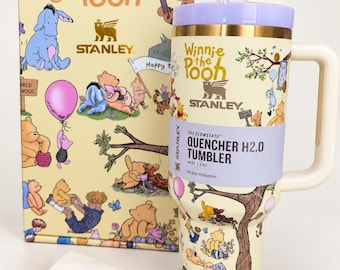 May include: A cream-colored Stanley Quencher H2.0 tumbler with a Winnie the Pooh print, a lavender lid, and a bee-shaped straw topper. The tumbler is displayed next to its matching illustrated box and a white fabric bag with Stanley branding.