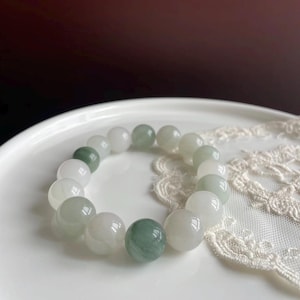 May include: A beaded bracelet composed of smooth, round, polished stones in varying shades of translucent white, pale green, and deep forest green. The beads are strung together in a circular arrangement, showcasing natural color variations.