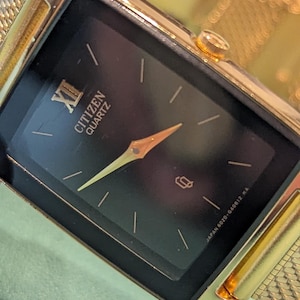 May include: A rectangular gold-tone Citizen Quartz wristwatch with a black dial. The face features a Roman numeral XII at the top, gold-colored hands, and simple stick indices for the remaining hours. The watch includes a gold-tone mesh metal band.