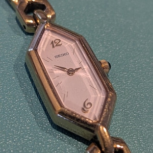 May include: A gold-tone Seiko wristwatch with a hexagonal case and a matching link bracelet. The watch face features a light-colored dial with the number 12 at the top, a small spiral design at the bottom, and thin gold-tone hands for timekeeping.