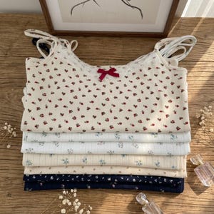 May include: A stack of five sleeveless camisole tops featuring thin shoulder straps and lace trim at the neckline. The top layer is cream with a red floral print and a small red bow. The other layers include white and navy blue with floral patterns.