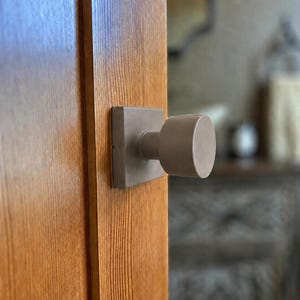May include: A matte taupe door knob with a square backplate mounted on a vertical wooden door panel. The knob features a cylindrical handle and a flat circular face, providing a clean and functional design for interior door hardware applications.
