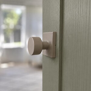May include: A matte beige cylindrical door knob with a square backplate mounted on a light sage green wooden door. The knob features a smooth, solid finish and a simple, functional design that sits flush against the flat surface of the door panel.