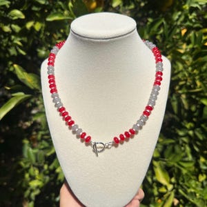 May include: A beaded necklace featuring alternating round red and translucent grey beads arranged in a repeating pattern. The necklace is secured with a silver-toned metal toggle clasp positioned at the front center of the strand.