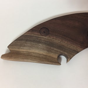 Op de afbeelding: Een houten serveerplank in de vorm van een vis. De plank is gemaakt van donkerbruin hout met een natuurlijk nerfpatroon. Het heeft twee cirkelvormige inkepingen en een gebogen uitsparing, waarschijnlijk voor een fles.