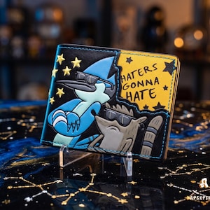 May include: A black leather bifold wallet featuring a graphic of a blue bird and a brown raccoon wearing sunglasses. The design includes yellow stars and the text "HATERS GONNA HATE" on a yellow background. The wallet has blue stitching along the edges.