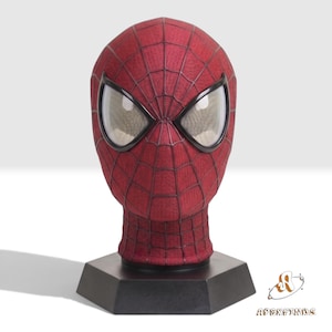 Puede incluir: Una máscara de tela roja con un patrón de telaraña negro y grandes lentes oculares de malla blanca, montada sobre un soporte de exhibición hexagonal negro. La máscara cubre toda la cabeza y el cuello, mostrando una textura detallada.