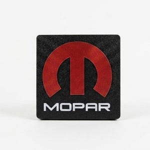 May include: A square black emblem featuring a prominent red Mopar logo centered on the front surface. The design includes the stylized letter M in red above the white text MOPAR, all set against a textured black background with rounded corners.