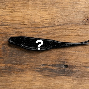May include: A black soft plastic fishing lure with a glittery finish and a split tail design. A large white question mark symbol is printed on the center of the lure body, which is shaped like a small baitfish with a tapered, pointed tail.