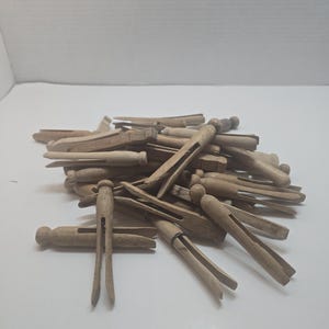May include: A pile of natural light brown wooden clothespins. Each clothespin features a classic two-piece design with a rounded head and a split body, showing the raw wood grain texture and color throughout the collection of multiple fasteners.