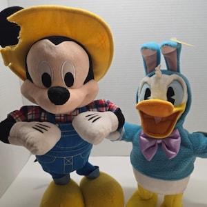 May include: A plush Mickey Mouse doll wearing a yellow hat, a red and white plaid shirt, and blue overalls, standing next to a plush Donald Duck doll wearing a blue hooded sweatshirt with bunny ears and a light purple bow tie.