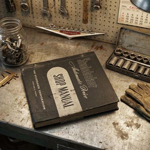 Könnte beinhalten: Ein schwarzes Studebaker Automatic Drive Werkstatthandbuch mit weißem Text auf dem Einband. Das Buch liegt auf einer Metallwerkbank neben einem Paar beigefarbener Lederarbeitshandschuhe, einem Messschieber, einem Glas mit Kleinteilen und einem Steckschlüsselsatz.