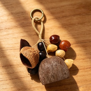 Puede incluir: Un llavero con un anillo de latón unido a un cordón beige, que sostiene un grupo de diversos elementos naturales, incluyendo semillas pulidas de color marrón oscuro, una cáscara de nuez texturizada y una pieza de madera redondeada.