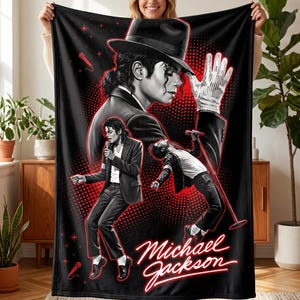 Puede incluir: Una manta de forro polar negra con una ilustración gráfica de Michael Jackson en varias poses, incluyendo el uso de un sombrero fedora y un guante de lentejuelas. El diseño incluye detalles en rojo neón, micrófonos y el nombre Michael Jackson en cursiva.
